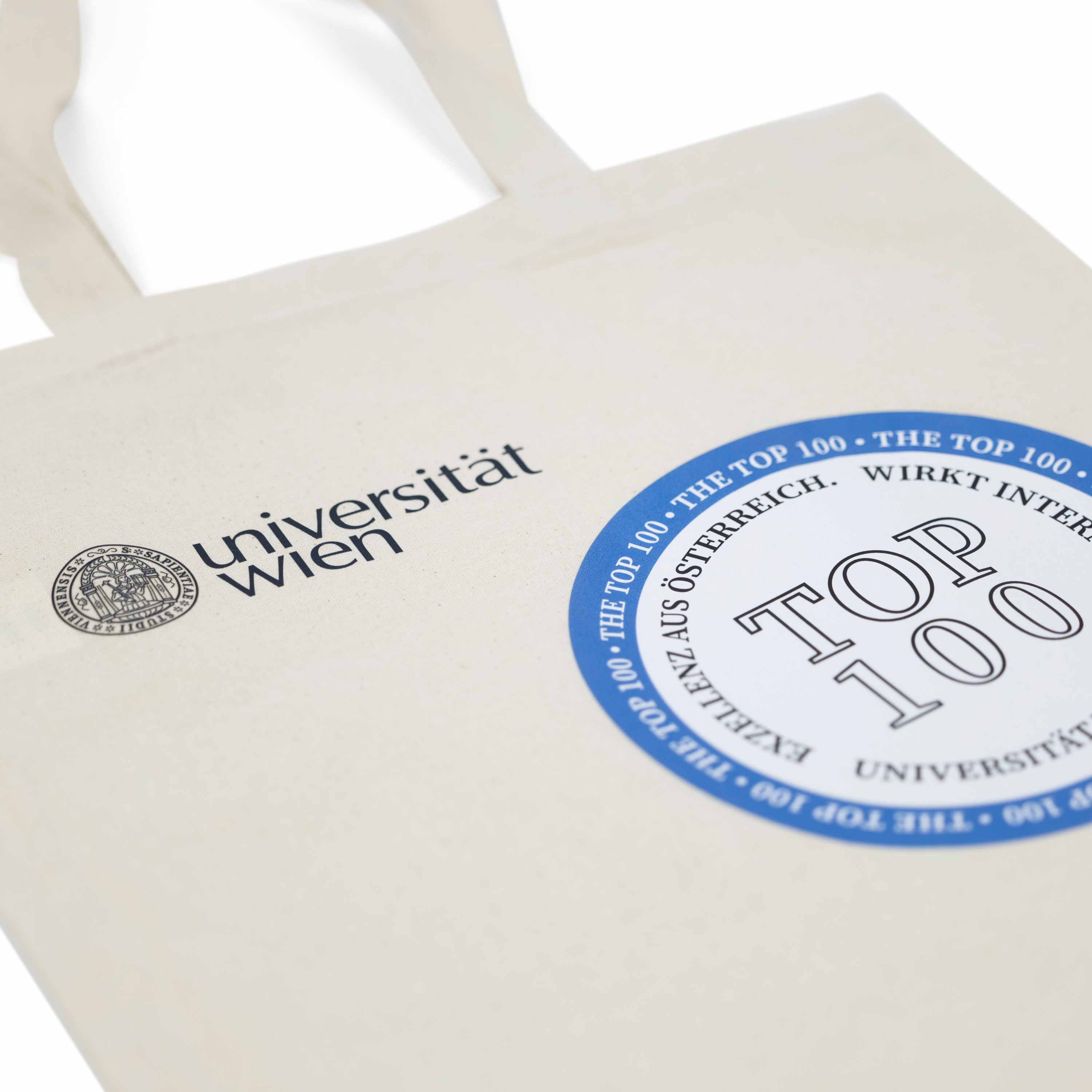 Nahaufnahme einer hellen Canvas‑Tasche mit Henkeln. Aufdruck links: Logo der Universität Wien. Rechts daneben ein blauer runder Badge mit der Aufschrift „TOP 100“.