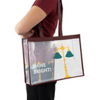 Transparente Tragetasche mit bordeauxfarbenem Rand und Schulterriemen; bedruckt mit gelb‑grünen Leselampen und dem weißen Schriftzug „SHINE BRIGHT!“.