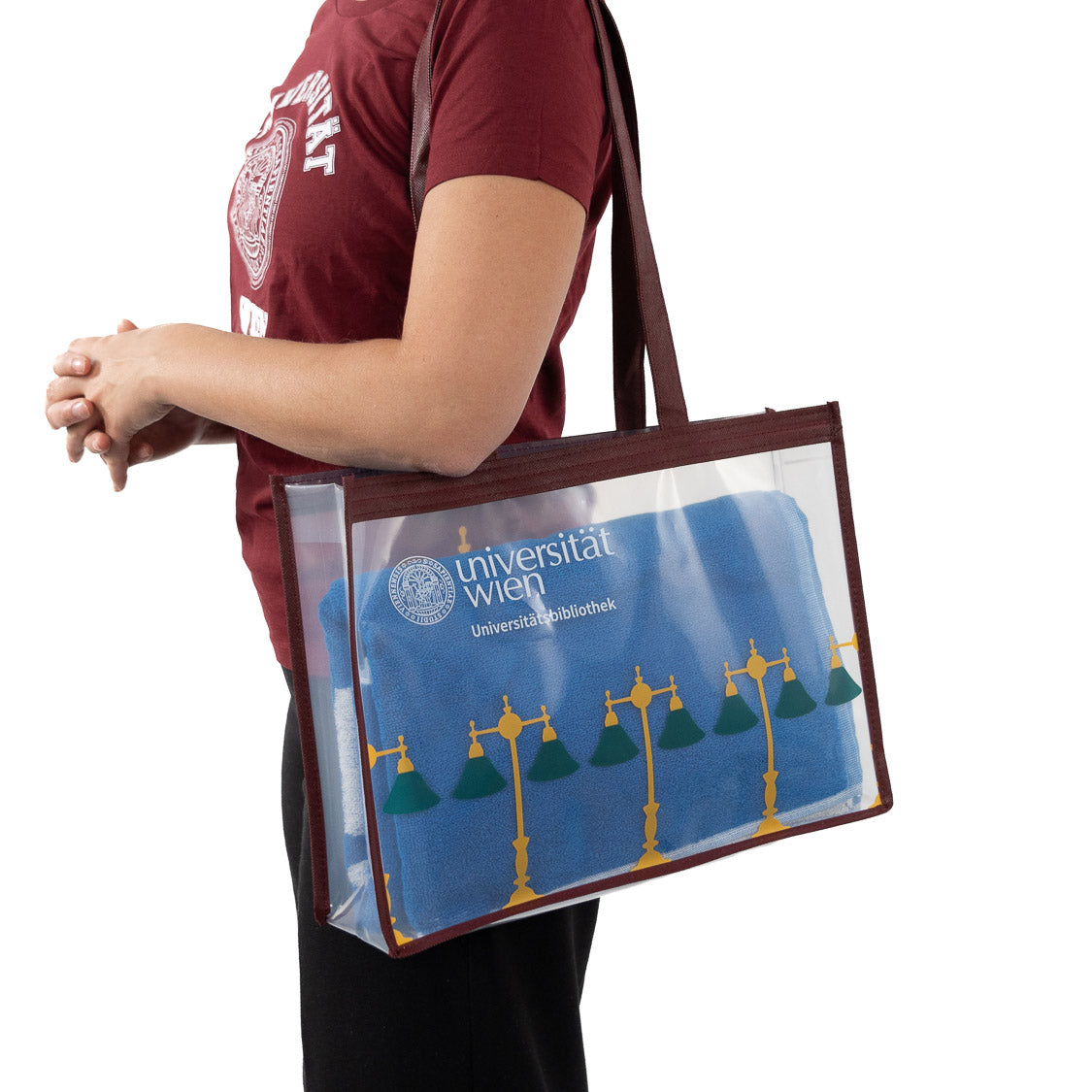 Transparente Tragetasche mit bordeauxfarbenem Rand und Schulterriemen; darauf das Logo „universität wien – Universitätsbibliothek“ und ein gelb‑grünes Lampenmotiv. In der Tasche liegt ein blaues Handtuch.