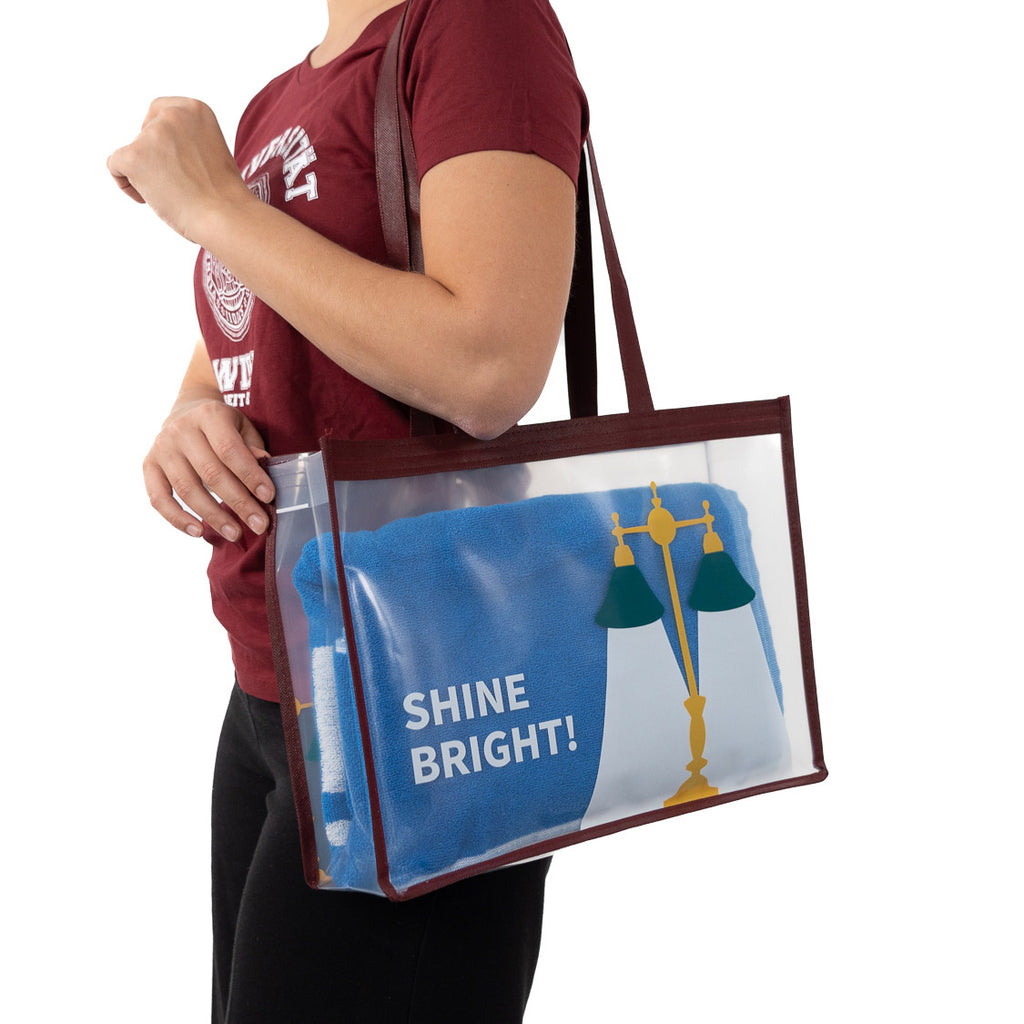 Transparente Tragetasche mit bordeauxfarbenem Rand, langen Henkeln und mit der Aufschrift „SHINE BRIGHT, getragen über der Schulter. In der Tasche liegt ein blaues Handtuch; außen ist ein gelb‑grünes Lampenmotiv zu sehen.